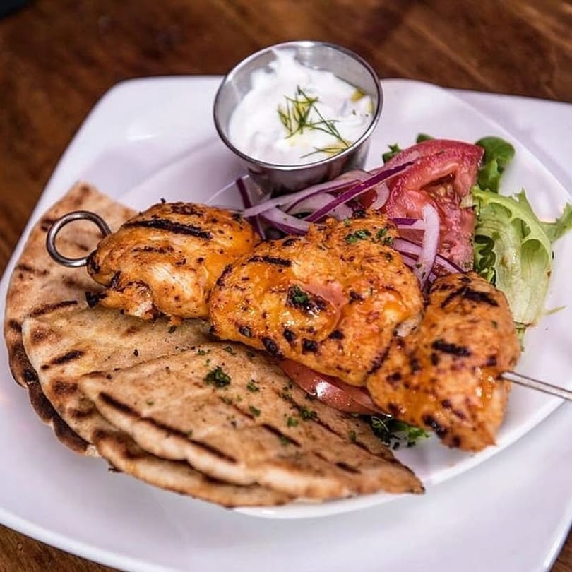 Grilled chicken souvlaki skewer with pita bread, tzatziki sauce, and a side salad on a white plate.