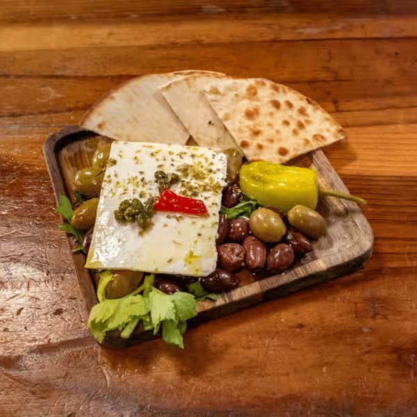 Feta cheese block with olives, capers, red pepper, and pita bread on a wooden board.