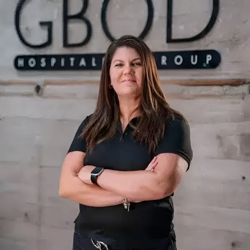 Woman with long brown hair and a black polo shirt stands with arms crossed in front of a sign.