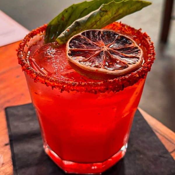 A vibrant red cocktail with a chili-lime rim, garnished with a dried lime slice and basil leaves.