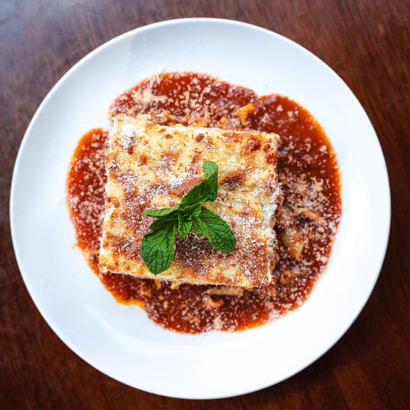 Golden-brown lasagna with red sauce, parmesan cheese, and a mint garnish on a white plate.