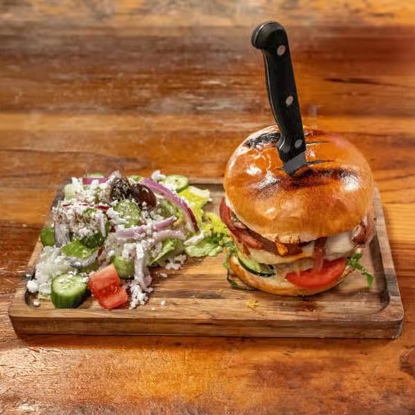 A large burger with a knife through it, served with a side salad on a wooden board.