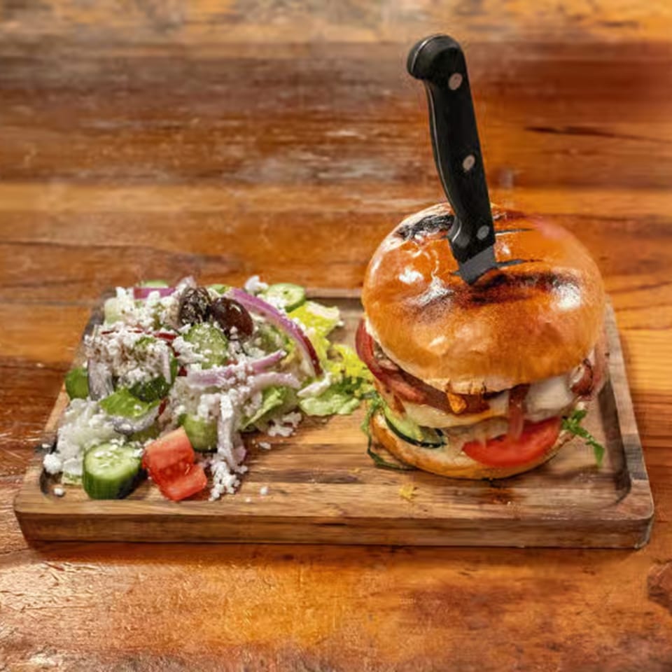 A large burger with a knife through it, served with a side salad on a wooden board.