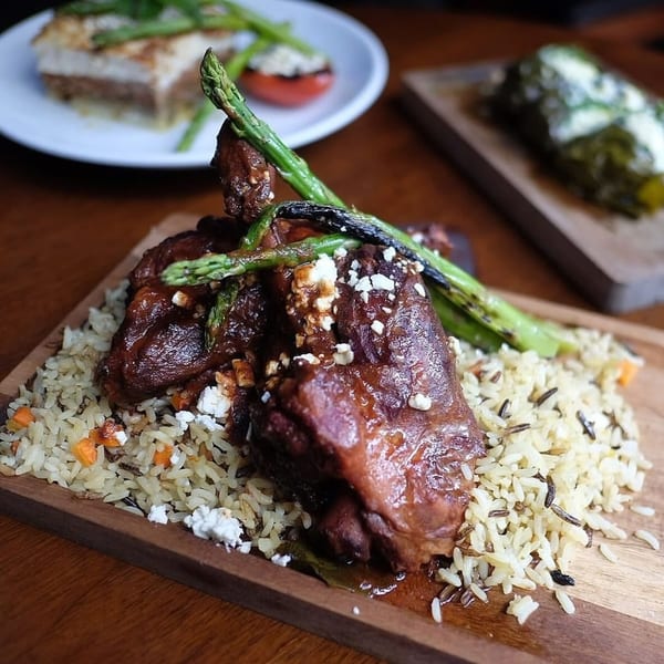 Braised lamb shank with rice pilaf, asparagus, and feta cheese on a wooden board.