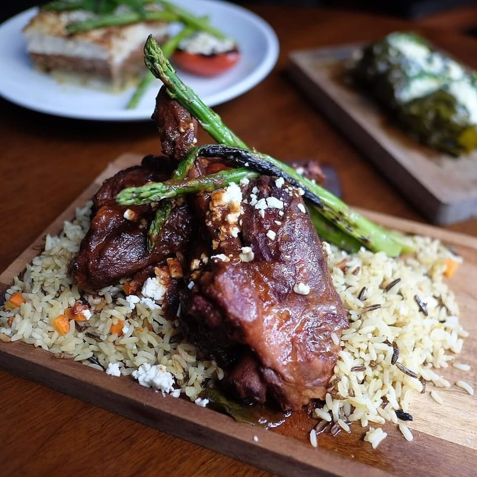 Braised lamb shank with rice pilaf, asparagus, and feta cheese on a wooden board.