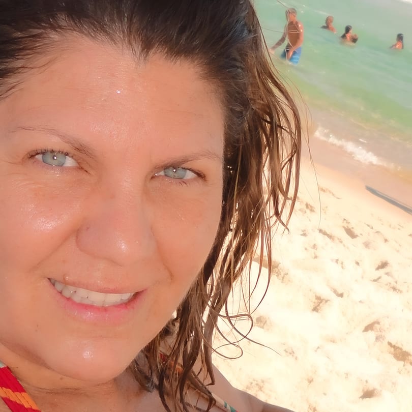A woman with wet hair smiles at the camera, with a beach and ocean in the background.