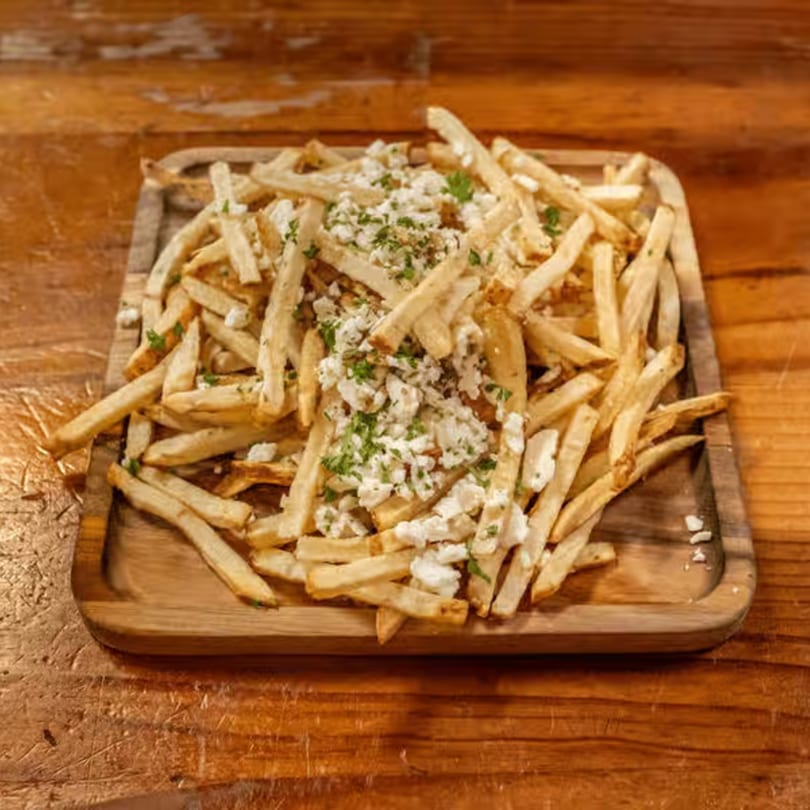 A wooden tray filled with golden french fries, topped with crumbled feta cheese and chopped parsley.