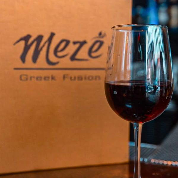 A glass of red wine in front of a menu board for Meze Greek Fusion restaurant.