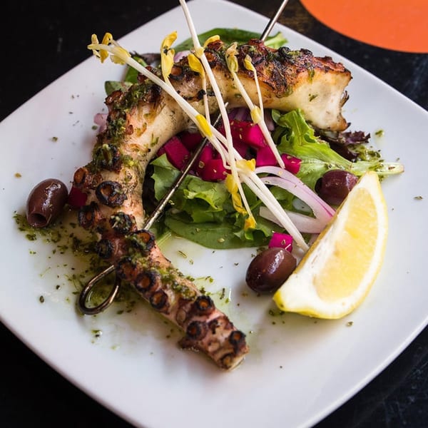 Grilled octopus with green herbs, olives, red onions, lettuce, and a lemon wedge on a white plate.