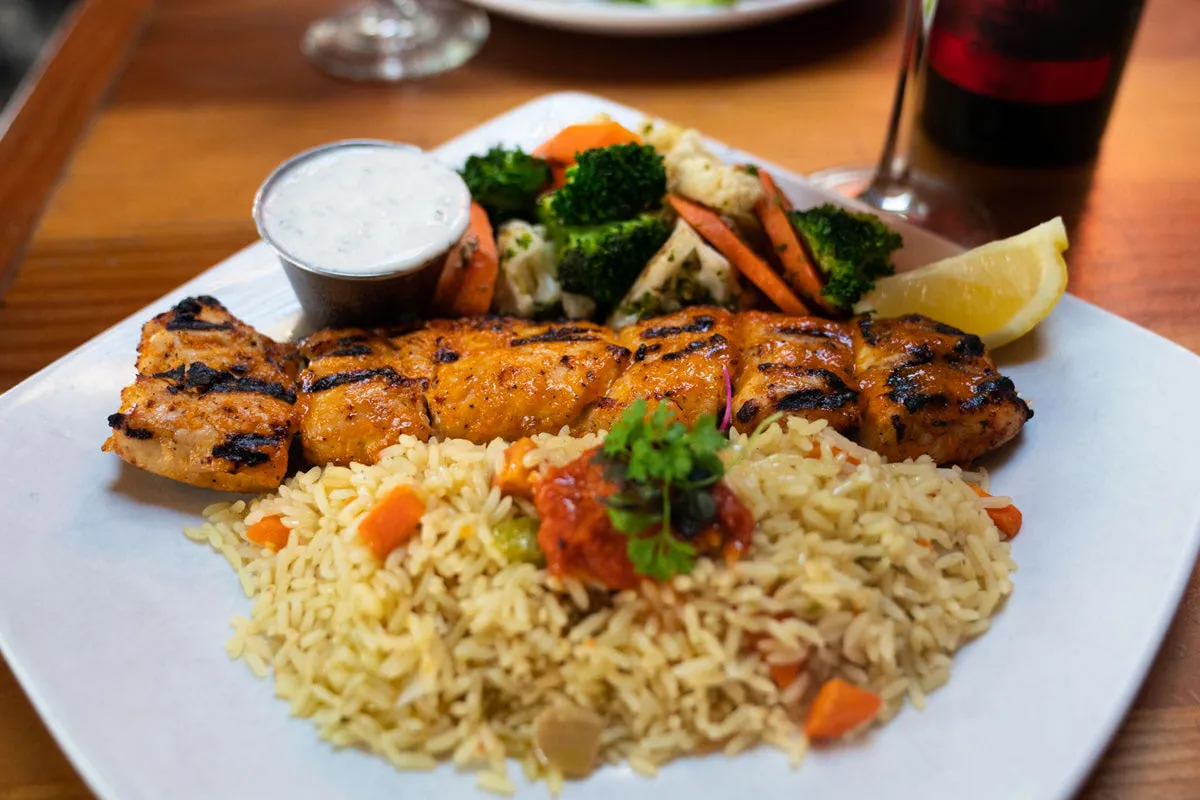 A photo of a plate of greek food, including rice, chicken kabob, and steamed vegetables